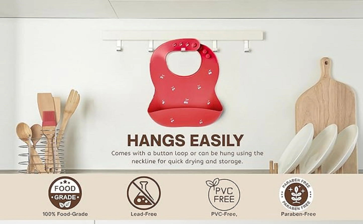 Red baby bib with cherry print hanging on a hook in a kitchen with wooden utensils and plates, food-grade, lead-free, PVC-free, paraben-free