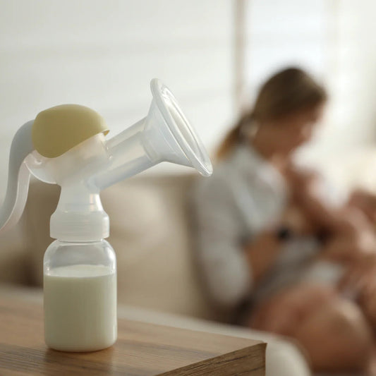 Manual breast pump with milk bottle on table, blurred mother breastfeeding baby in background