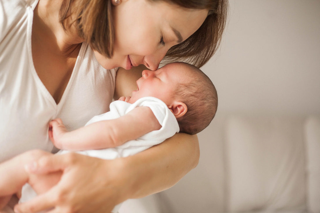 Mother lovingly holding and nuzzling newborn baby wrapped in white cloth on neutral background